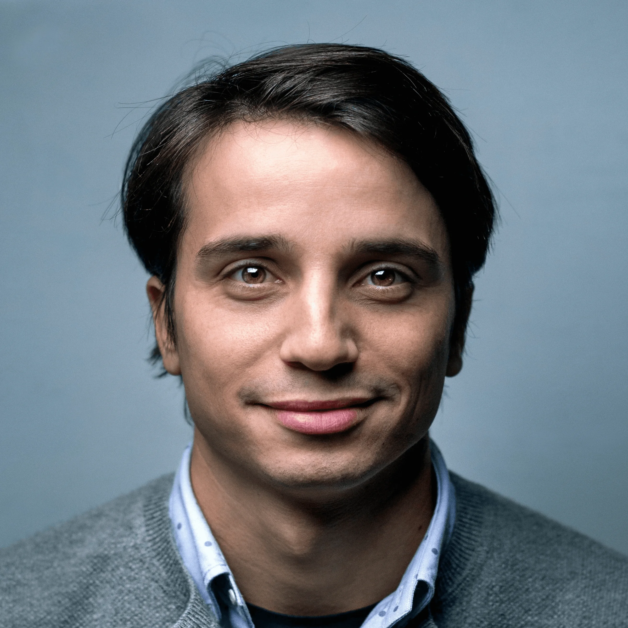 Person in grey sweater and blue shirt facing camera with neutral expression against light grey background
