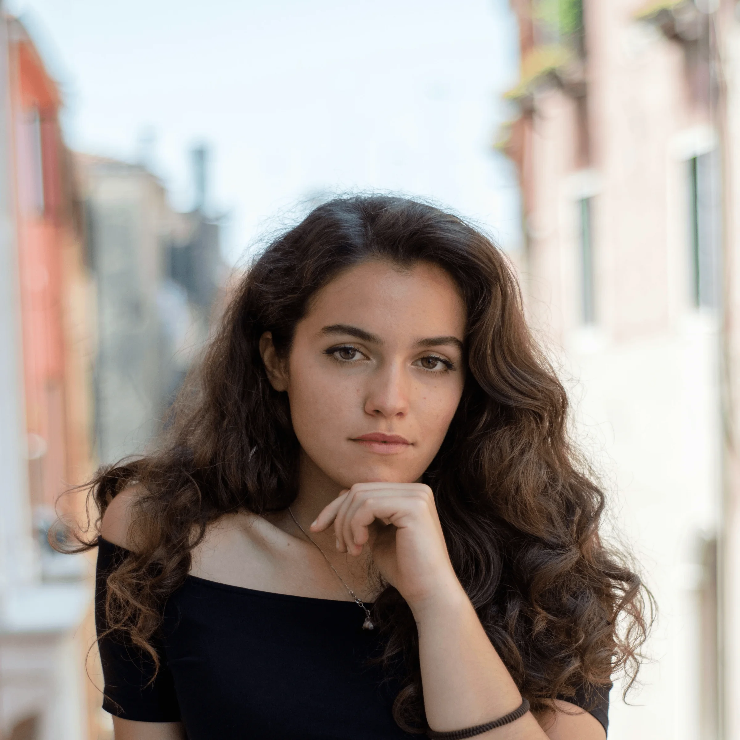 Person in black top posing with hand under chin in urban alleyway, blurred buildings behind