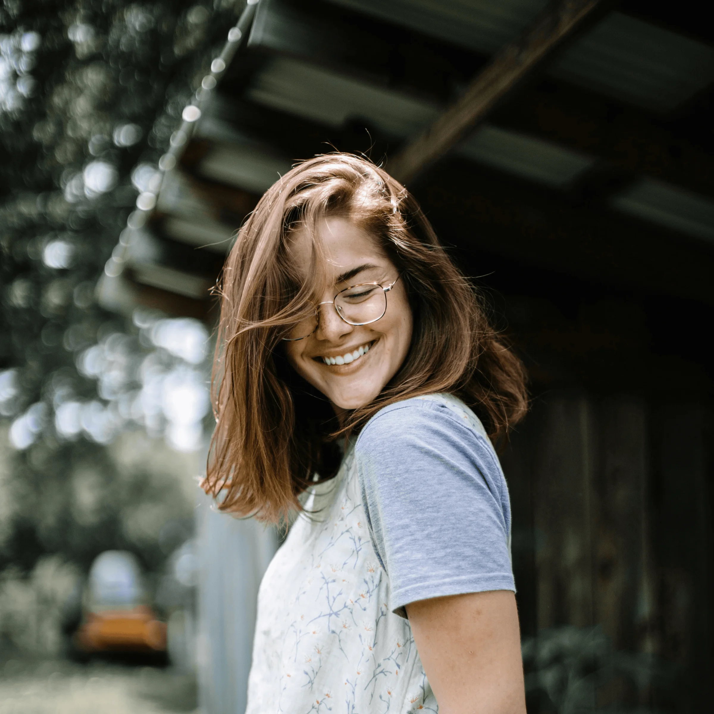 Person in floral shirt with glasses standing outdoors near wooden shed and yellow vehicle.