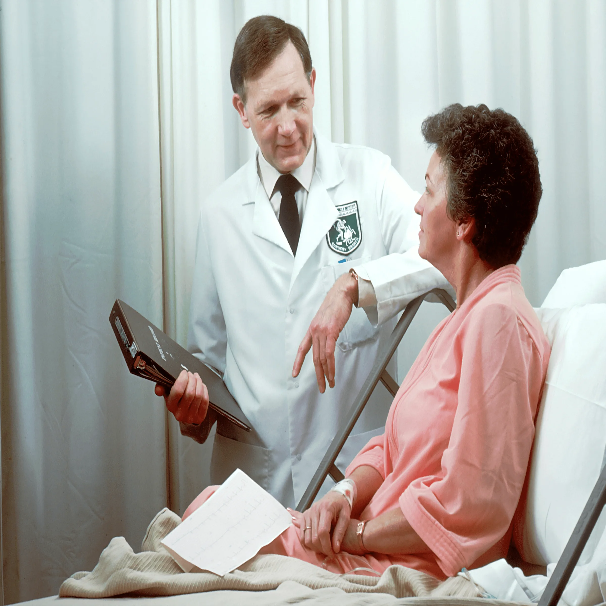 Doctor in white coat discussing ECG results with patient in pink gown in hospital room.