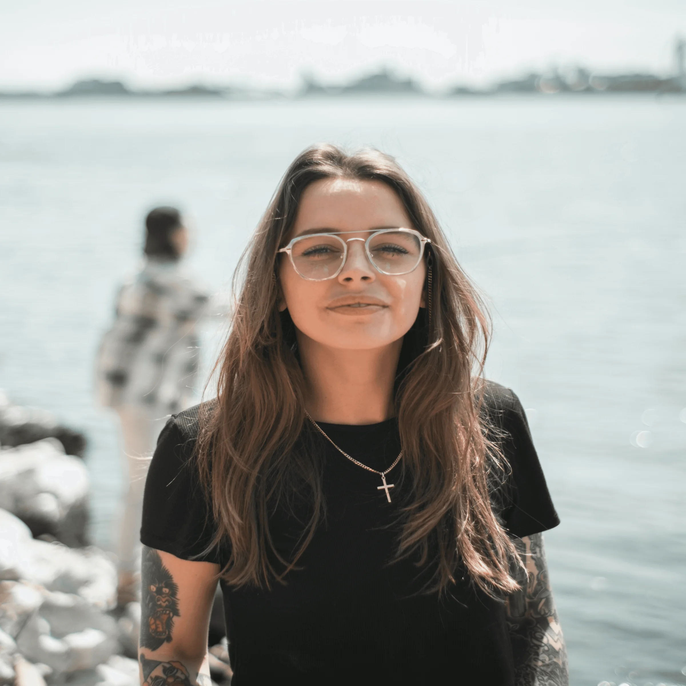 Person with tattoos and cross necklace standing by water, another figure on rocks in background.