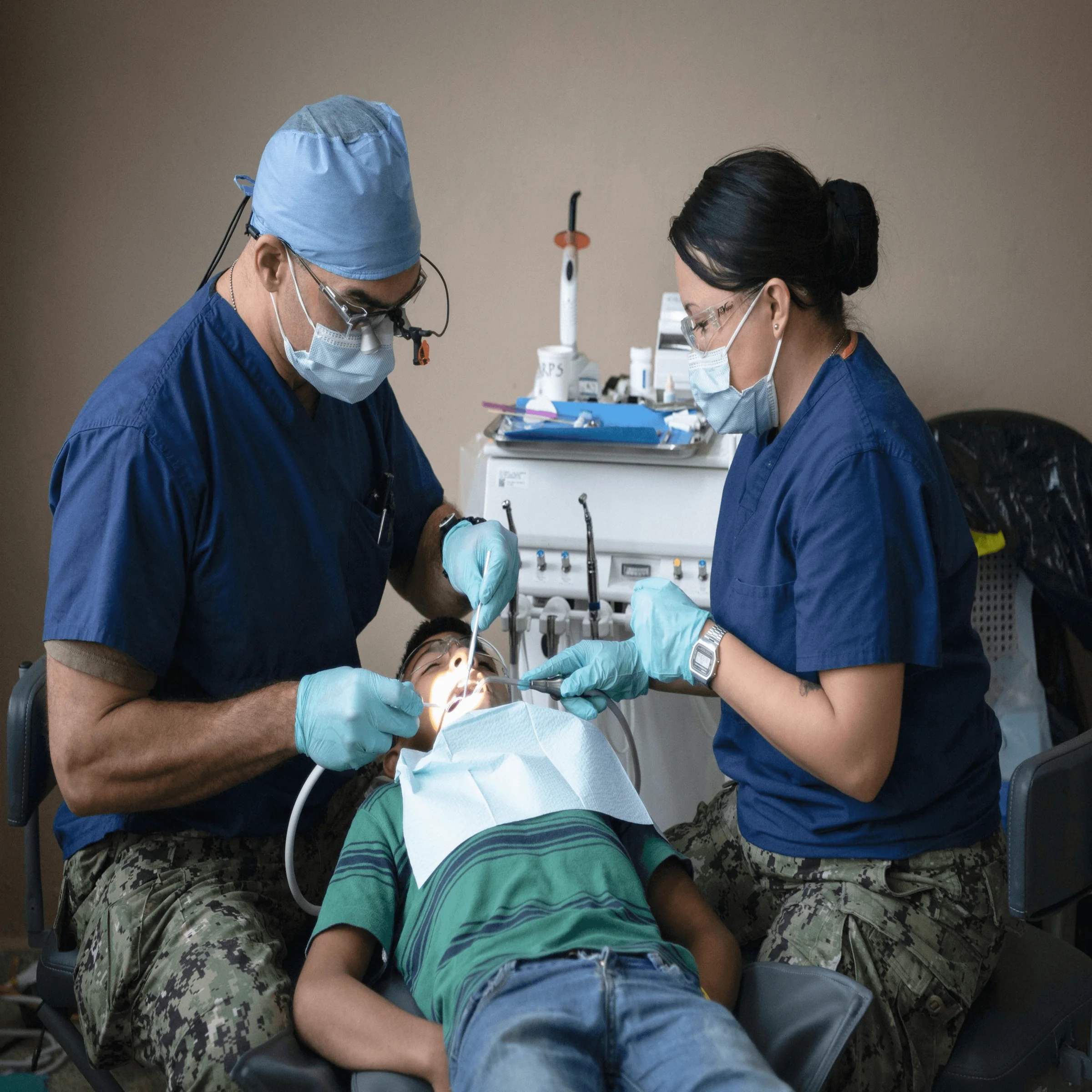 Two dental professionals in blue scrubs treating young patient in clinic with tools and equipment