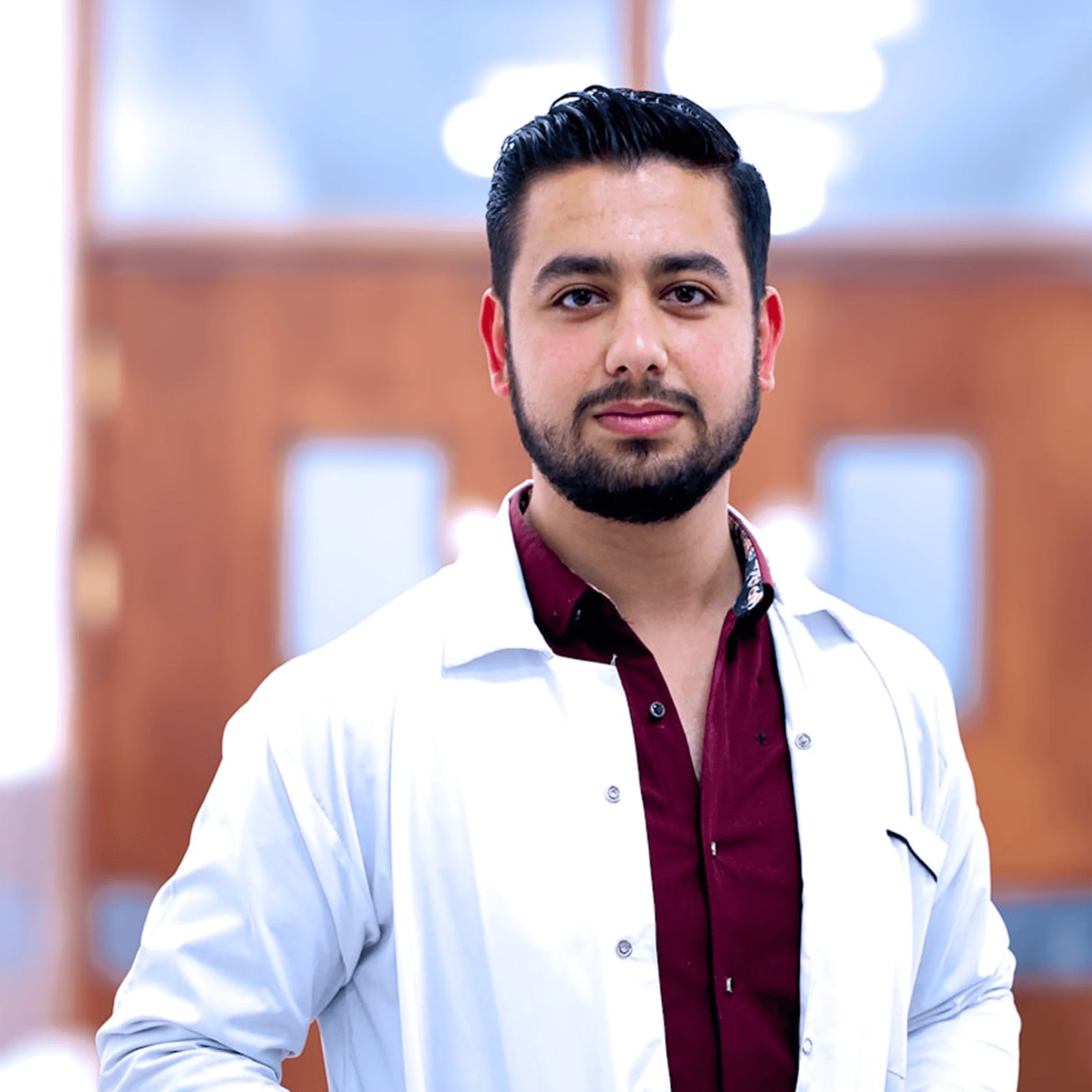 Person in white lab coat over maroon shirt standing in hallway