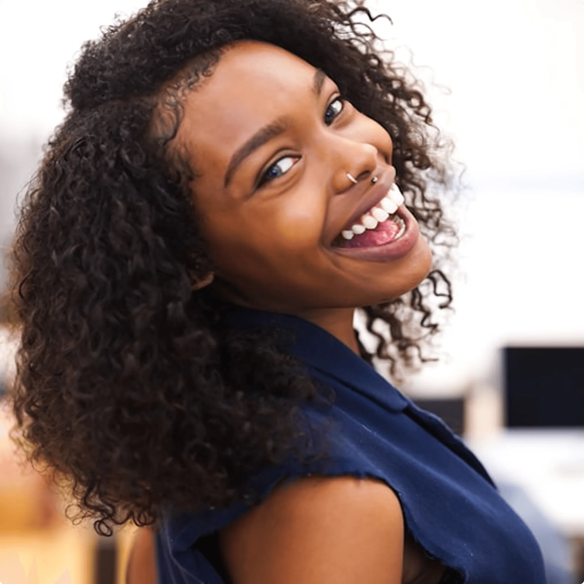 Smiling person with curly hair and nose ring looking over shoulder in bright indoor setting.